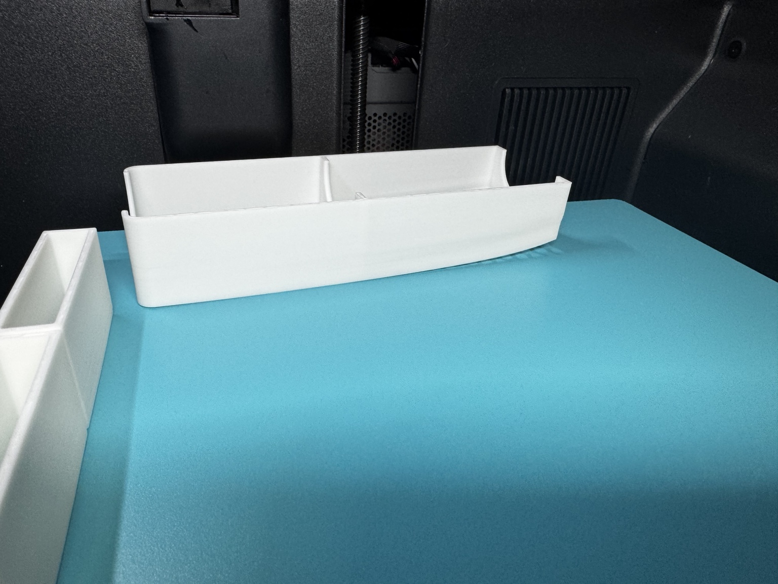 Close view of a long white organiser with its right-hand end lifted above the blue build plate.